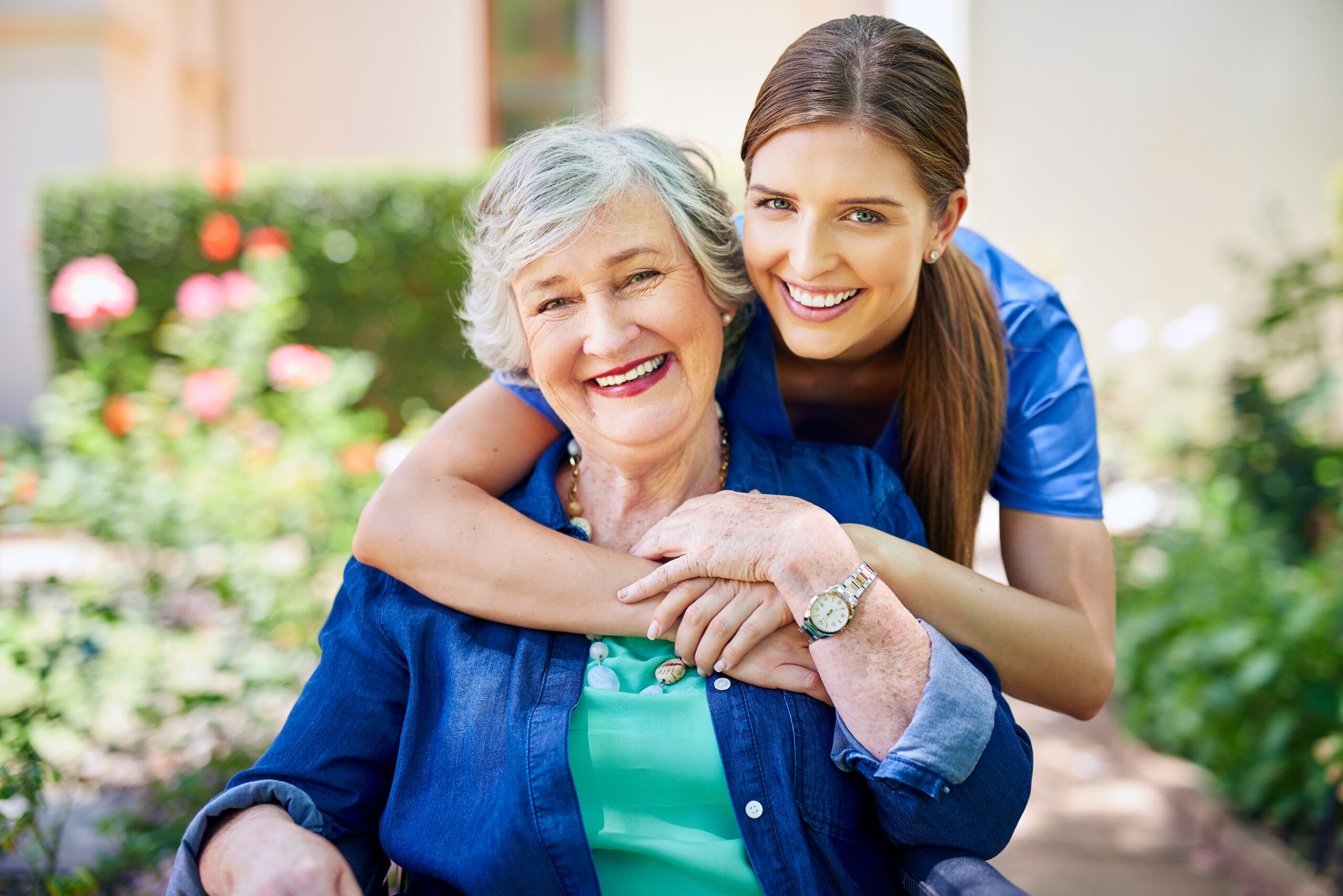 A young woman embraces an elderly woman outdoors, both smiling warmly.