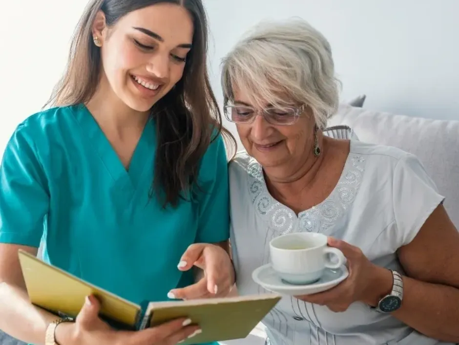 A nurse showing a tablet to a smiling elderly woman drinking tea.