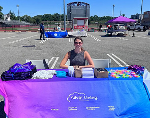 A woman sits behind a table with Silver Lining branded items at an outdoor event.