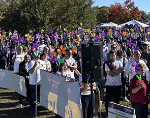 A crowd gathered outdoors, many wearing purple, enjoying a sunny event with speakers and tents.