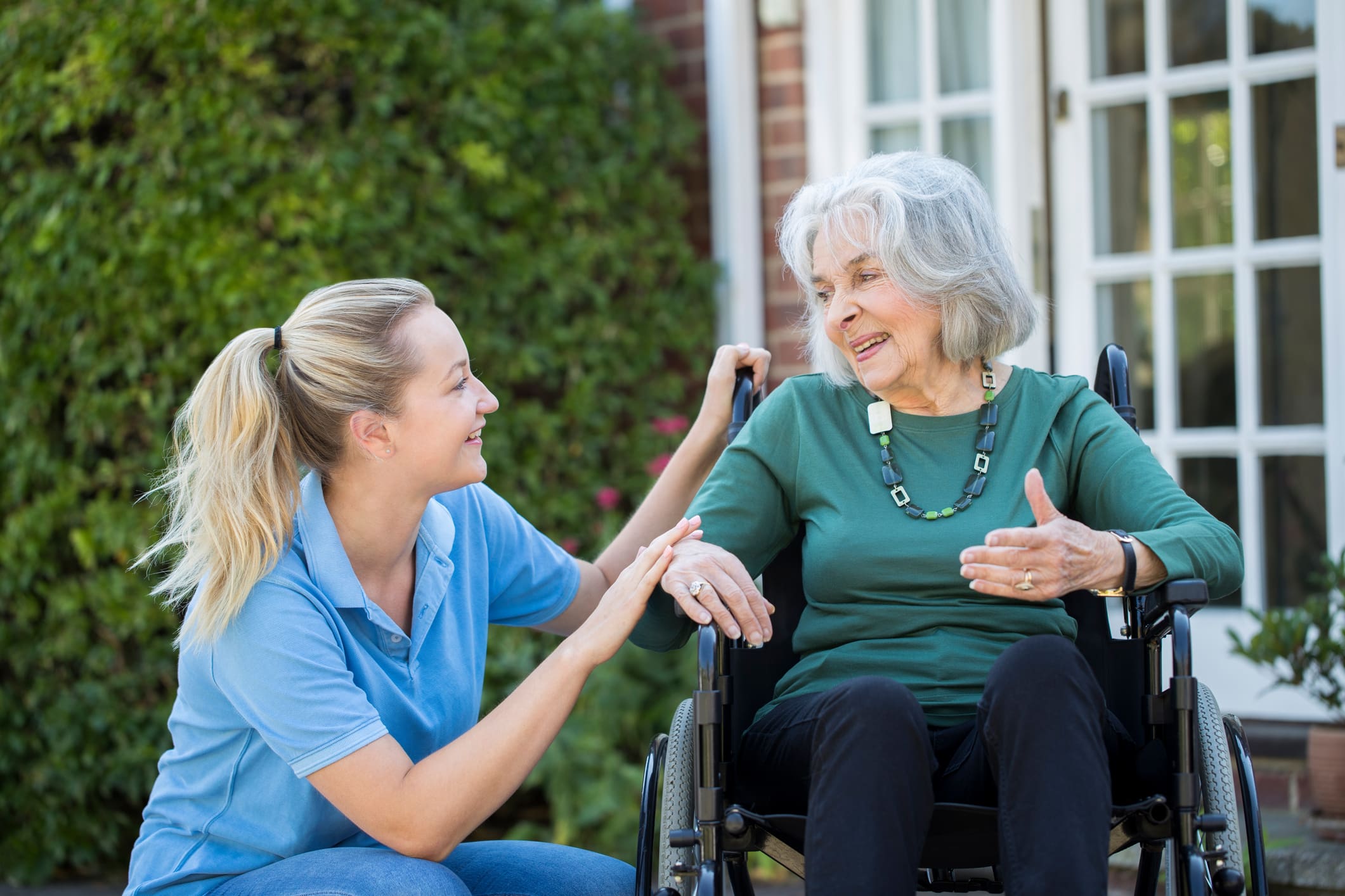 Caregiver smiling and chatting with elderly woman in wheelchair outdoors.