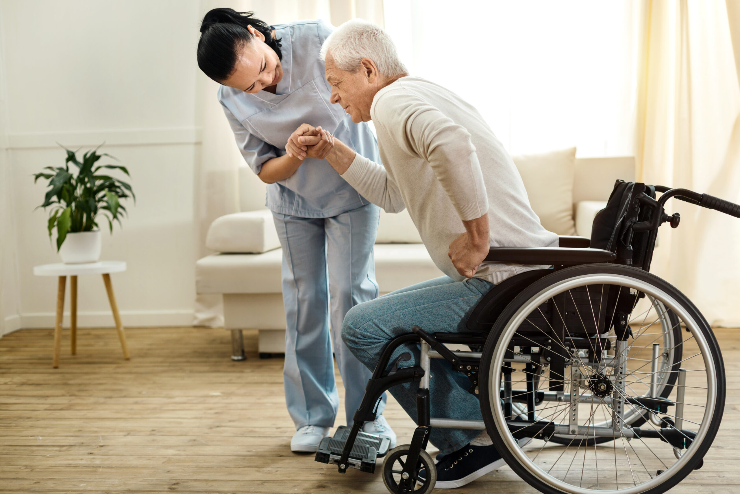 A caregiver helps an elderly man in a wheelchair stand up.